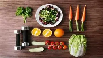 mesa de madeira com cenoura, laranja, tomate, abobrinha e halteres