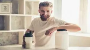 Homem com barba na cozinha sorrindo com shake na mão e apoiando o outro braço em cima do pote de suplemento