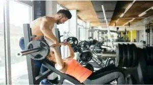Benefícios da musculação para saúde, instrutor ajudando a aluna na acadmeia a fazer exercício de levantamento de peso para o peitoral