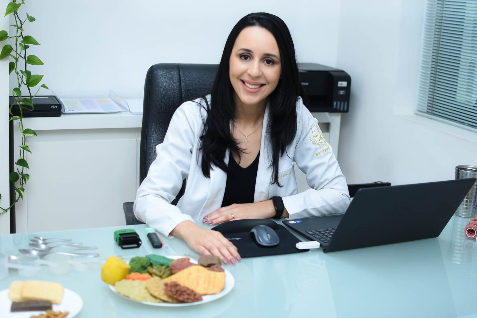 Tainá Carvalho sorrindo em sua mesa de atendimento com um prato de alimentos saudáveis